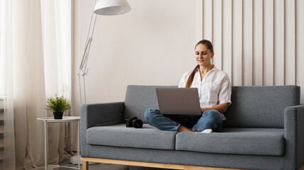 Happy young woman using laptop at home. On the couch next to the woman is a camera. Photographer...