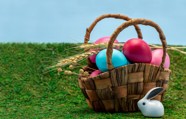 Colorful Easter eggs in a basket and a small figure of an Easter bunny on the green grass. Free space. Easter composition, bright color, festive mood.