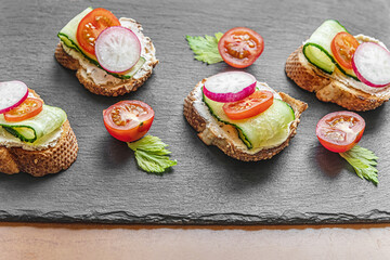 toasted baguette slices with cream cheese, cucumber, radish and cherry tomatos on a black slate board