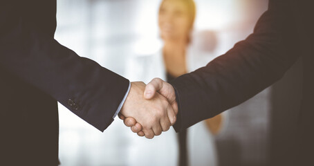 Unknown businesspeople are shaking their hands after signing a contract, while standing together in a sunny modern office, close-up. Business communication, handshake, and marketing concept