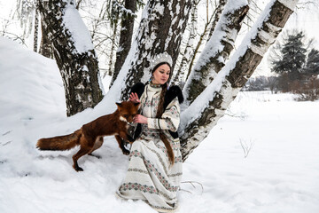 Russian beauty in a national costume with a red fox in a winter snowy forest 