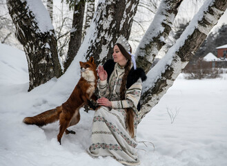 Russian beauty in a national costume with a red fox in a winter snowy forest 