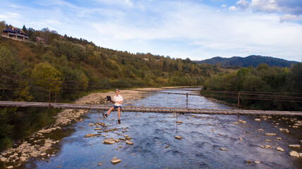 Aerial drone view of young man in white shirt sitting on wooden bridge over mountains river and using laptop. Success freelancer 