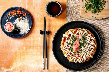 Homemade japanese fast food okonomiyaki cabbage pancake decorated by spring onion, pickled ginger, mayo sauce on black ceramic plate with chopsticks and ingredients above. Texture background. Flat lay - 413880415