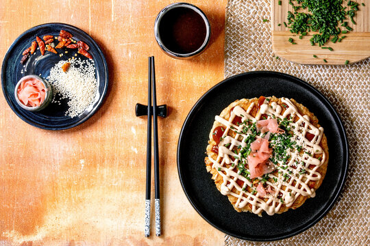 Homemade japanese fast food okonomiyaki cabbage pancake decorated by spring onion, pickled ginger, mayo sauce on black ceramic plate with chopsticks and ingredients above. Texture background. Flat lay