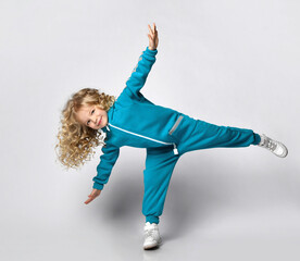 Smiling happy girl in warm fashion jumpsuit trendy sportswear fooling around standing like airplane at studio copy space. Child beauty model with blond curly hair advertising sportive clothing