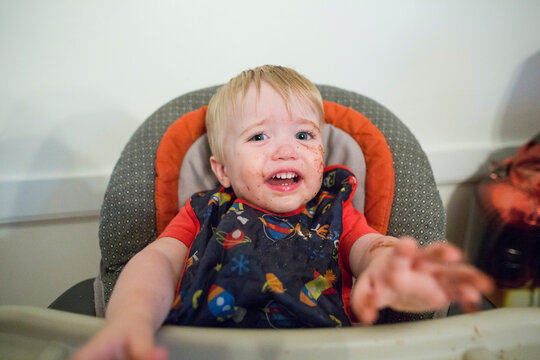 Portrait of messy baby boy crying while sitting on high chair against wall at home