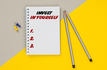 On a yellow-gray background are pencils, buttons and a notebook in which it is written - INVEST IN YOURSELF