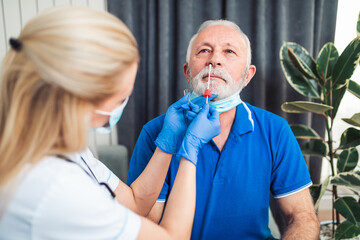 Female doctor visiting examining senior male patient at his home. He is taking sample from throat and nose of man suspected to be Covid-19 positive.