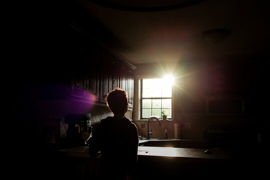 Silhouette boy looking bright sun through window while standing in kitchen