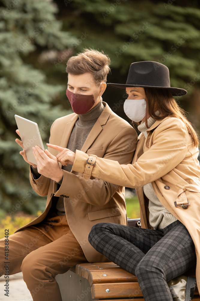 Poster young stylish couple in protective masks using tablet while resting in park