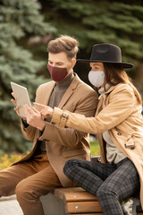 Young stylish couple in protective masks using tablet while resting in park