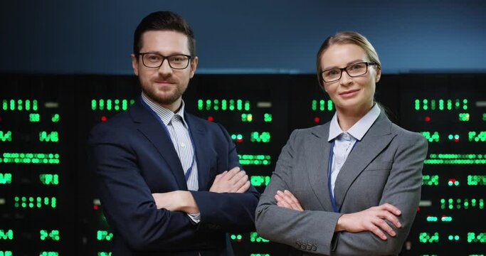 Portrait of two smart and successful programmers, man and woman, standing on background of running servers looking at camera with arms crossed.