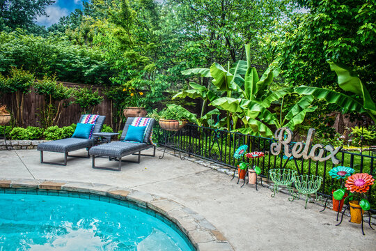 Two lounge chairs by swimming pool on concrete deck with tropical plants and metal scultures and sign that says Relax.
