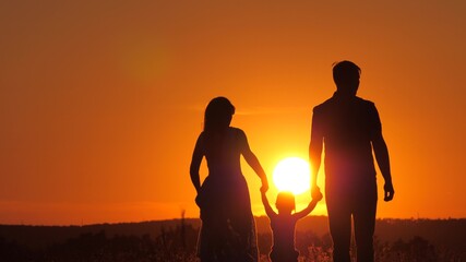 Fototapeta na wymiar Happy family, little daughter is jumping, holding hands of dad and mom in park in sun. The child plays with dad and mom on field in light of sunset. Walk with small child in nature. childhood
