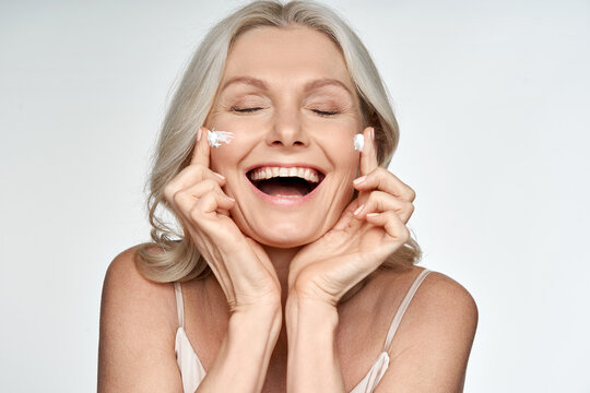 Happy Funny 50s Middle Aged Mature Old Blond Woman Applying Facial Cream On Face Laughing Enjoying Skin Care Beauty Spa Treatment Creme Isolated On White Background. Mid Age Skincare Concept.