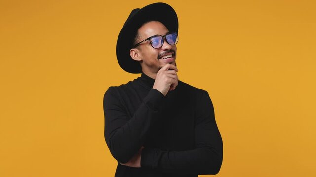 Smiling pensive young african american man 20s in casual black clothes hat eyeglasses isolated on yellow color background in studio. People lifestyle concept. Looking aside up put hand prop up on chin