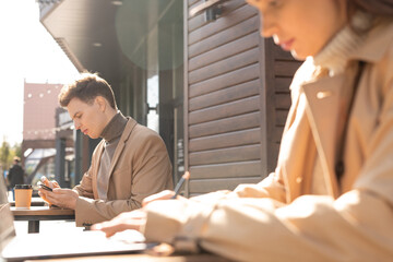 Side view of young elegant man in jacket and sweater using smartphone outdoors