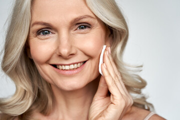 Smiling 50s middle aged older woman holding cotton pad sponge cleansing face skin with cleanser,...