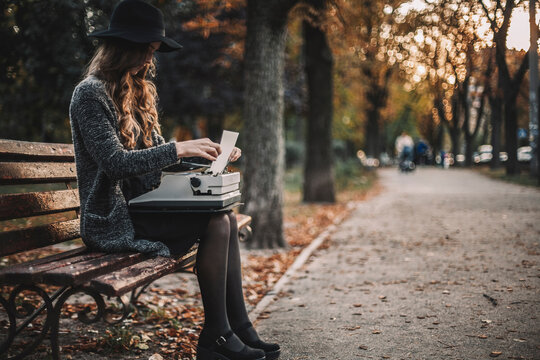 Side view of female writer using typewriter while sitting on bench at park