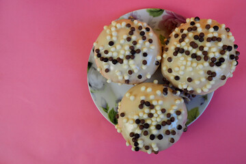 close up three round doughnuts in white frosting with chocolate balls in a white plate with flowers on a pink background top view