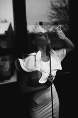 Black white photo portrait of beautiful model in white dress touching transparent glass keeping plant