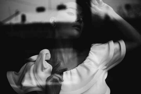 Black white photo portrait of beautiful woman in white dress posing near transparent glass wearing make-up