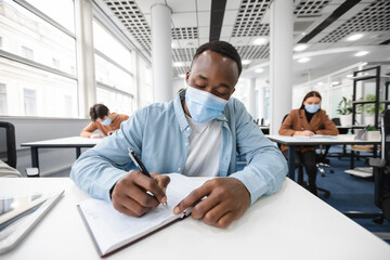 Black guy in medical mask sitting at desk and writing