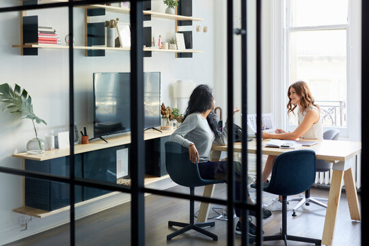 Female colleagues working together while sitting at desk in office seen through window