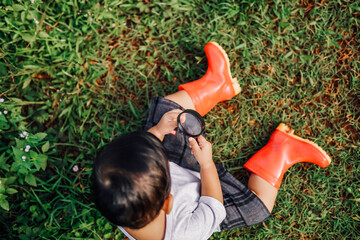 a happy kid sits on the grass wearing a red rubber boot carrying a magnifying glass