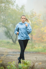 Fototapeta na wymiar Blond sportswoman jogging along forest path among trees on foggy morning