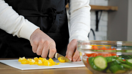 Close up hands professional chef cook man in black apron standing near table sliced fresh yellow pepper on cutting board at home kitchen. Preparing ingredients food vegetables for vegetarian salad