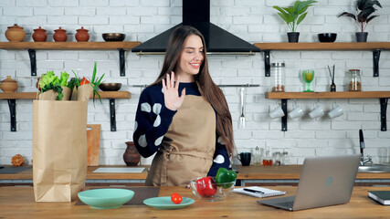 Smiling woman housewife in beige apron greets waves hand with teacher study online remote video call conference webcam laptop computer, distance education chef culinary course school at home kitchen