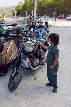 child boy in a green jumpsuit walks among old motorcycles in the summer