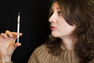 Obraz premium Vaccine concept. Female portrait of caucasian girl holding a syringe and looking at it on black background in studio.