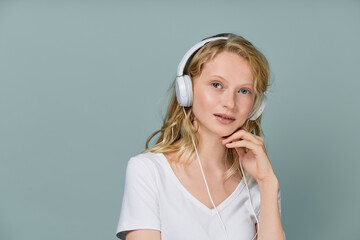 Closeup portrait of young woman closed eyes listening music via headphones on color neutral gray tone wall. Pretty serious clever blonde woman with curly hair looking at camera. Vertical