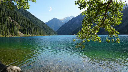 Mountain lake Kolsay with floating boats. View of green hills, fir trees, grass and water. Boats with tourists float on the lake. People are resting. The water of a mountain lake is green.