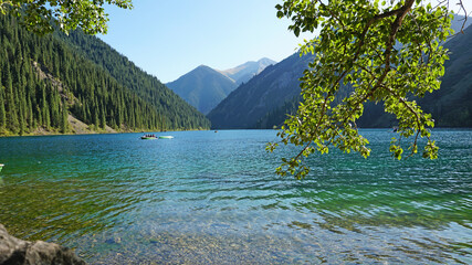 Mountain lake Kolsay with floating boats. View of green hills, fir trees, grass and water. Boats with tourists float on the lake. People are resting. The water of a mountain lake is green.