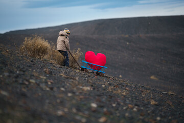 A determined child, despite the difficulties, pulls a stroller with a large plush heart up the hill...