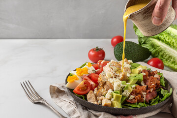 A hand pouring salad dressing into cobb salad with chicken, avocado, bacon, blue cheese, tomato and...