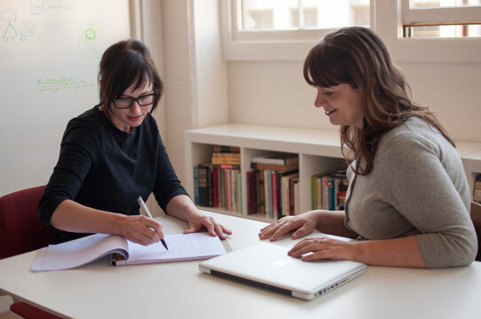 Businesswoman looking at female colleague writing on book in office