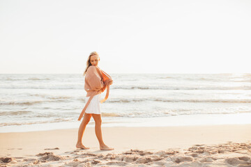 Fototapeta na wymiar Young slim woman walk alone on the beach or ocean at summer sunset. Female dressed in a warm sweater.