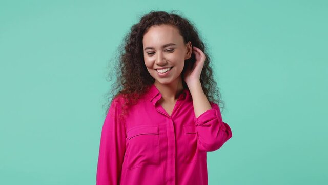 Smiling cheerful young african american woman 20s years old in pink shirt posing isolated on blue turquoise background in studio. People sincere emotions lifestyle concept. Looking camera blinking