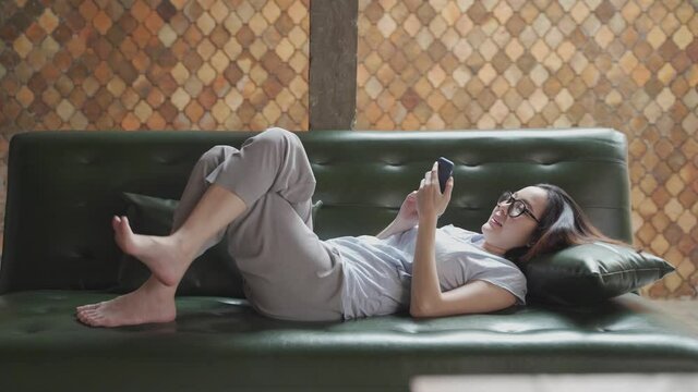 happy asian woman relaxing using mobile smart phone surfing internet and video call on vintage sofa couch at home. selective focus on phone.