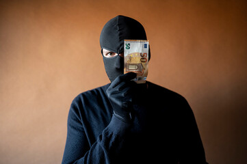 Male thief with balaclava on his head holding a handful of euro bills in front of his eyes