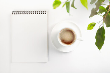 Mockup top view of an open blank notebook on a spiral,, white porcelain cup with coffee and leaves of a houseplant. Selective focus, soft focus. Copy space. White background.