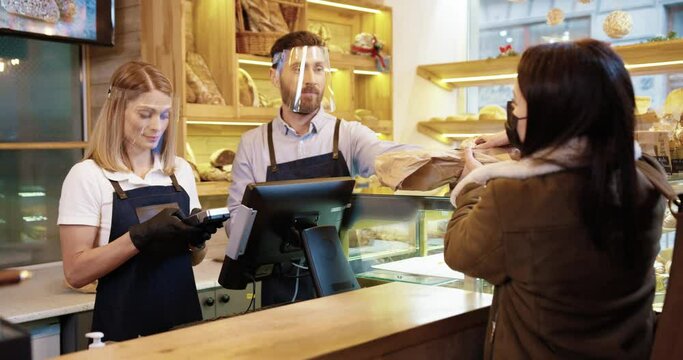 Portrait of Caucasian young couple of bakers sellers in face shields and gloves working selling fresh bread to young female customer standing in small bakehouse. Rear of woman paying with credit card