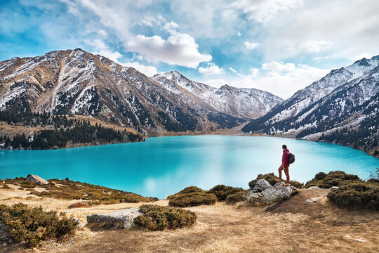 Full length of backpacker looking at view while standing by lake against mountains and cloudy sky during winter