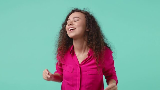Funny cheerful young african american woman 20s in basic pink shirt isolated on blue turquoise background studio. People lifestyle concept. Dancing clenching fists waving rising hands snapping fingers