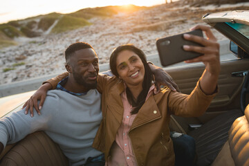 Diverse couple sitting in a convertible car and taking a selfie